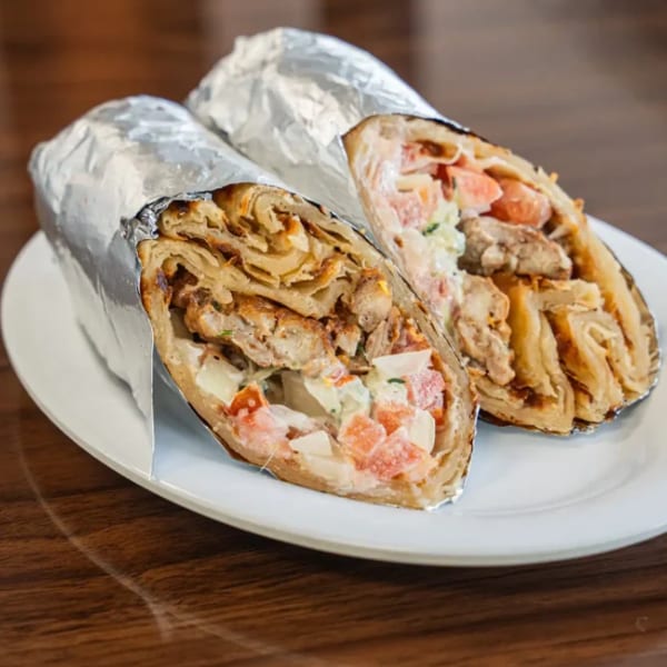 Two foil-wrapped shawarma wraps, cut in half, revealing chicken, tomatoes, and sauce on a white plate.
