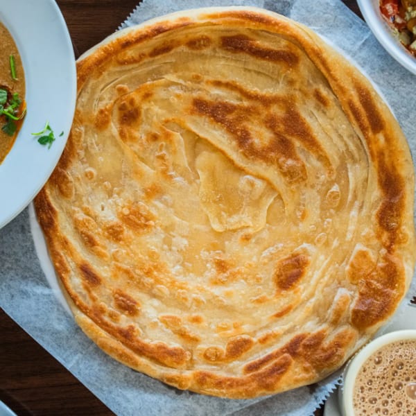 Golden, flaky paratha bread on a white plate, surrounded by various Indian dishes and a cup of chai.