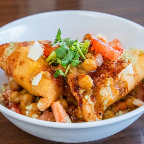 Samosa chaat with chickpeas, tomatoes, and cilantro in a white bowl on a wooden table.