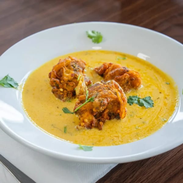 Three pakoras in a yellow curry sauce, garnished with cilantro, on a white plate with a spoon.